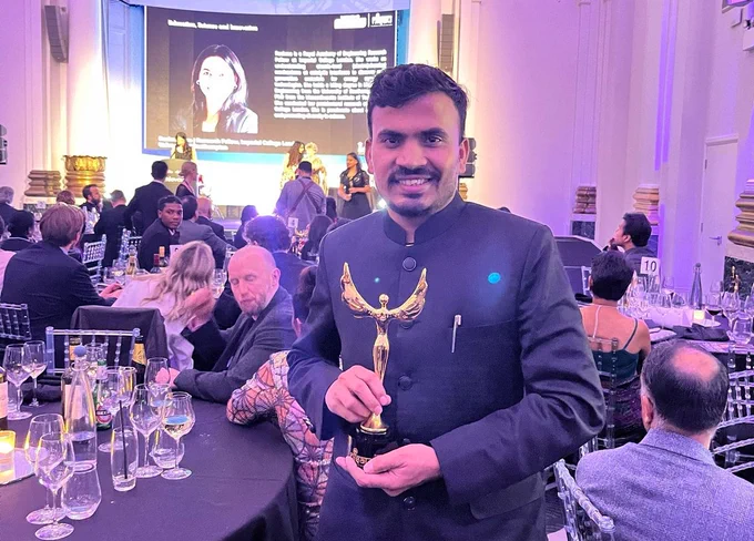 Person holding a trophy at an award ceremony with attendees seated at tables and a presentation screen in the background displaying text and an image of a woman.