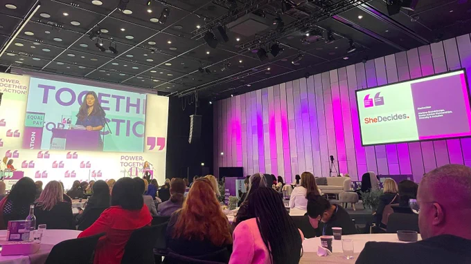 Person speaking at a podium with "SheDecides" logo displayed on a screen in a conference room filled with attendees.
