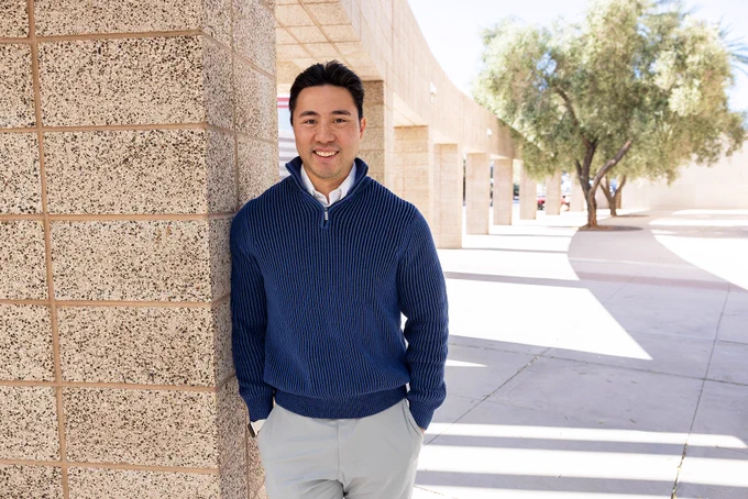 Man in blue sweater standing against a stone wall with architectural structures and trees in the background.