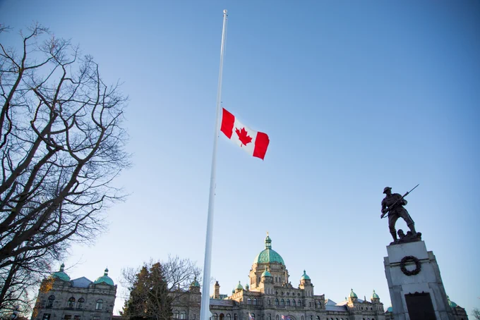 Canadian flag flying at half-mast against a blue sky with Parliament Buildings in the background