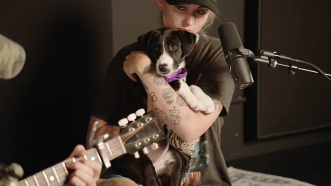 a woman in a black t-shirt holds up a black and white puppy against her chest. her arm is covered in small black and white tattoos.