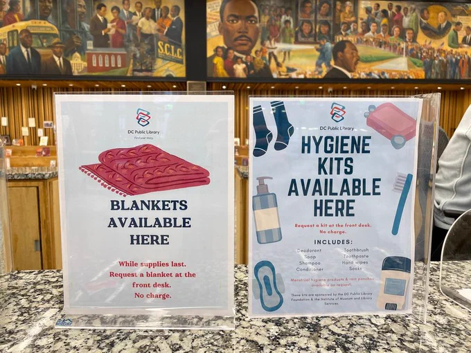 Two signs placed on a counter at Dr. Martin Luther King Jr. Library. Left sign advertises "Blankets Available Here" with a request to ask at the front desk. Right sign offers "Hygiene Kits Available Here," listing items included like shampoo and toothbrush. Each sign features the library's logo. A colorful mural in the background depicts civil rights themes.