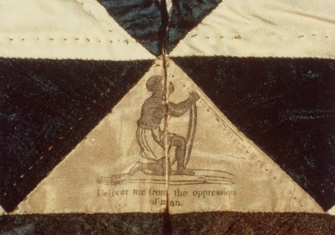 Close up photo of a cream and white patchwork fabric item, made up of alternating triangles that meet in the upper center of the image. Printed or stamped on the bottom cream triangle is a Black figure in chains, with text reading "Deliver me from the oppression of man."