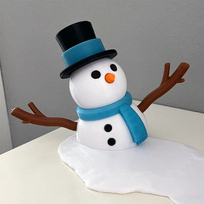 Photo of a 3D printed snowman "melting" on a table. The snowman is wearing a light blue scarf and a black top hat with a light blue band.