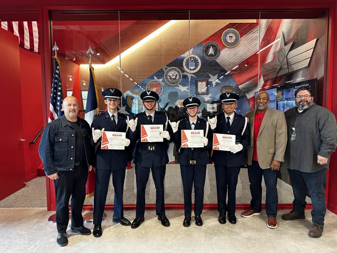 Group of seven individuals, including four in military uniform, holding certificates in front of a display with various military insignias at a formal ceremony. Two civilians stand on the left and right ends of the group.