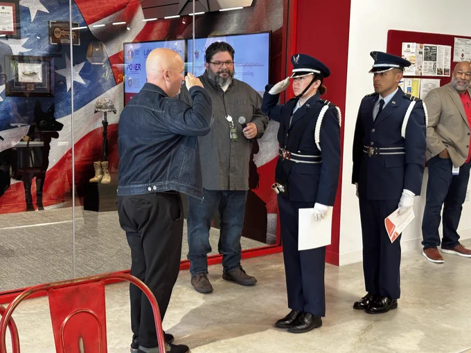Two individuals are engaged in conversation while exchanging a small object, with another person looking on nearby. They are flanked by two uniformed officers, standing at attention against a backdrop featuring a large American flag. Several onlookers observe the scene, which appears to be in a public, indoor setting decorated with patriotic themes.
