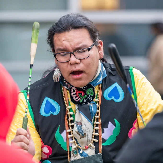 A person wearing native dress beats a drum and sings outdoors
