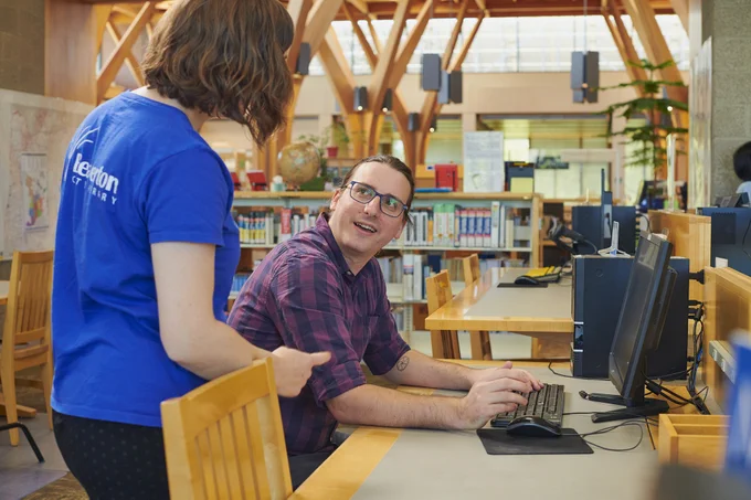 Photograph of a man at a computer in the library looking up at a library employee standing next to him. 