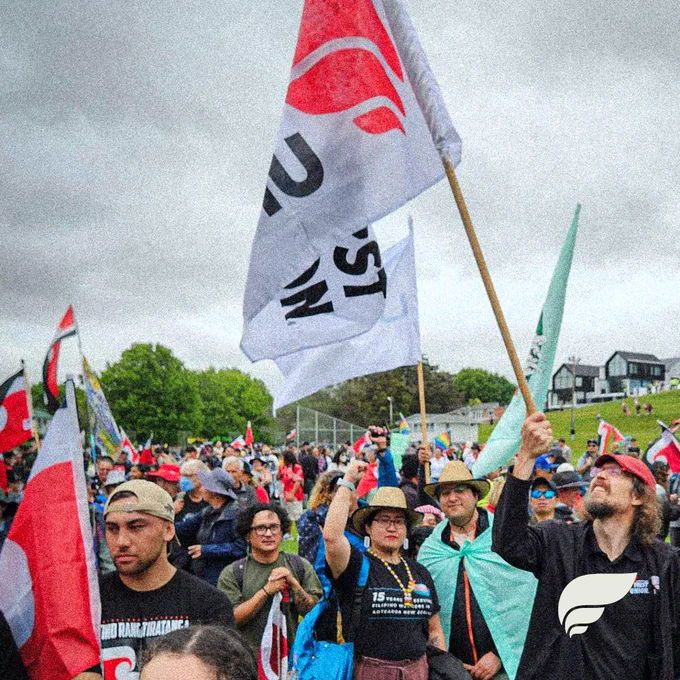 A group of people marching. A lot of Māori flags are visible. In the foreground a person holds a FIRST Union flag. A person just behind them is holding up their closed fist.