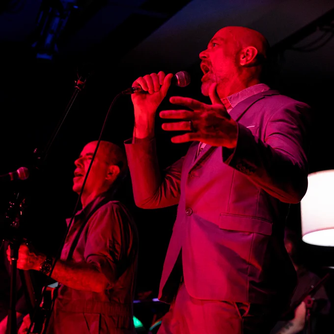 Two people in suits performing in front of microphones with red stage lighting.