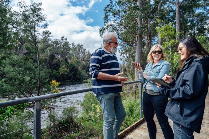 Three people standing in a forest next to a waterway, one person is taking notes