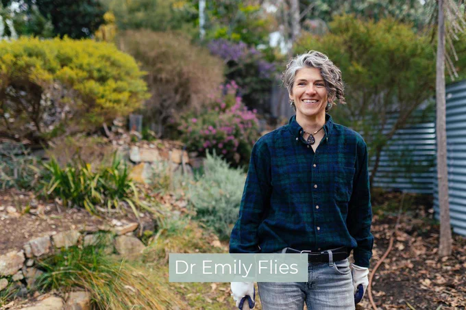 Dr Emily Flies standing in a garden, smiling and wearing gardening gloves