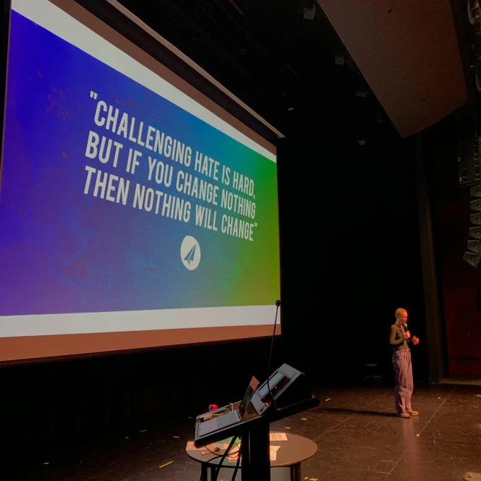 [ID: A photo of a presenter at a PROJECT ROCKIT workshop with a quote slide on the projector screen reading “Challenging hate is hard, but if you change nothing, then nothing will change.”]