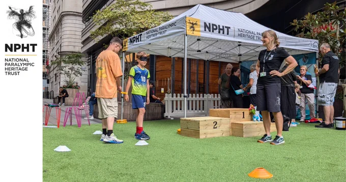 Learning Manager Fiona oversees one of our Incredible Me challenges. there are two boys, one is blindfolded and tethered to the other. There are several cones on ethe floor marking out a course for them to navigate. In the background is the NPHT pop-up museum, and a small wooden podium. 