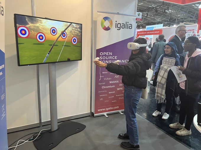 On the right, a person playing an archery game at the Igalia stand using VR glasses with a Wolvic browser sticker, while other people observe amused. On the left, a large screen next to them displays the game being played.