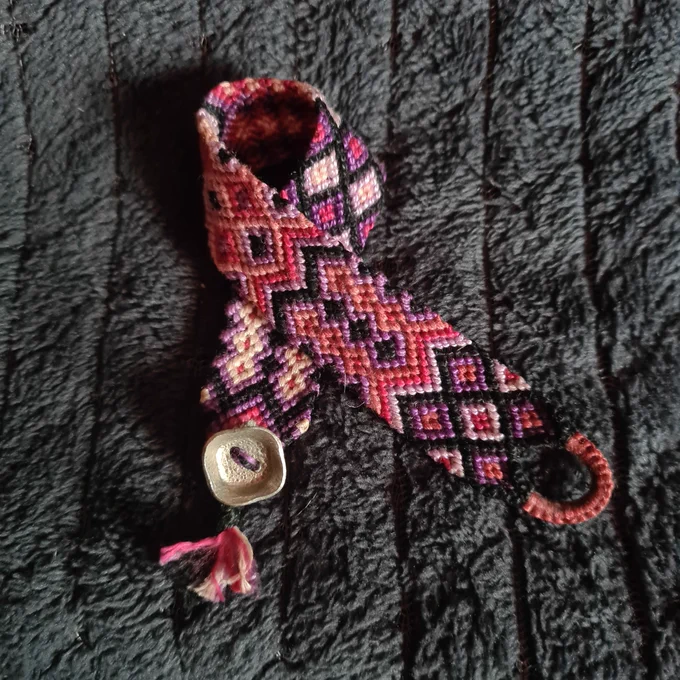 Un bracelet en macramé avec plein de formes géométriques dans les tons rose, parme, violet, noir, avec un bouton de métal argenté carré aux bords arrondis...