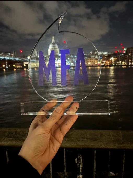 A glass trophy with purple letters - MFA (media freedom awards) is held up by Barbara’s hand. Through the glass, you can see the Thames and St. Pauls. The night sky is visible above with the lights of the city reflecting on the river.