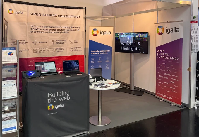 Igalia's booth at Electronica Fair 2024 showing a couple of tables with demo devices on top, including a set of VR glasses, several banners, a TV and a brochure stand. The devices also include a Steam deck and an ARM laptop.