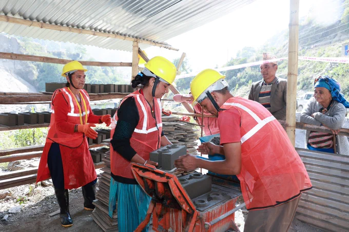 Two women and a man wearing red jackets and yellow helmets for safety making bricks made of climate friendly locally available materials. 