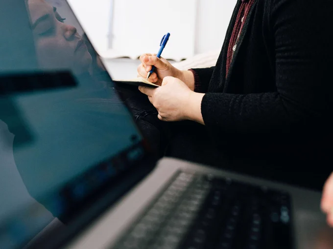 Close up of a laptop screen. In the background a person is holding a pen and paper