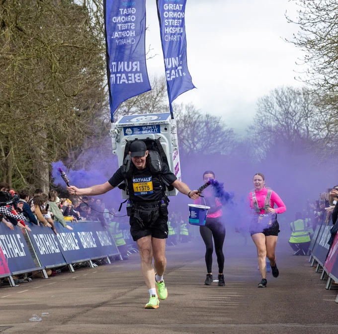 A photo of a man running down the final straight of the 2024 Bath Half with a heavy box with Great Ormond Street Hospital charity flags and labels attached. He is also holding two purple smoke bombs.