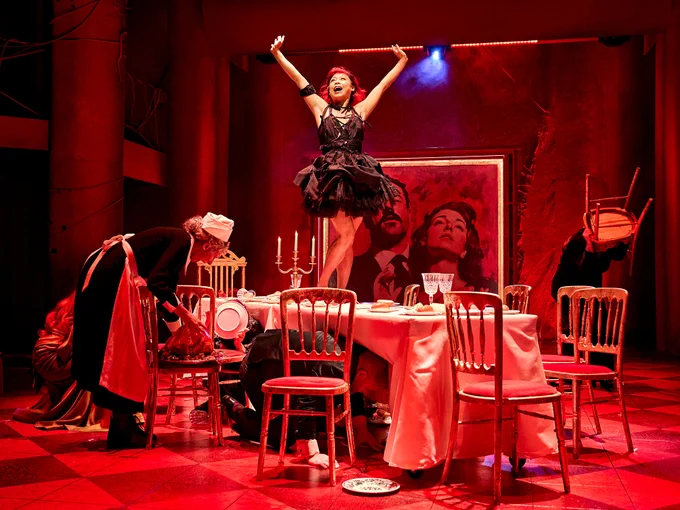Nikki Cheung as Karen dancing on a large table, which has been hastily tidied after a large meal. The lighting is intensely red, and Nikki wears a black dress with ruffled skirt. Other members of the company are tidying the table.