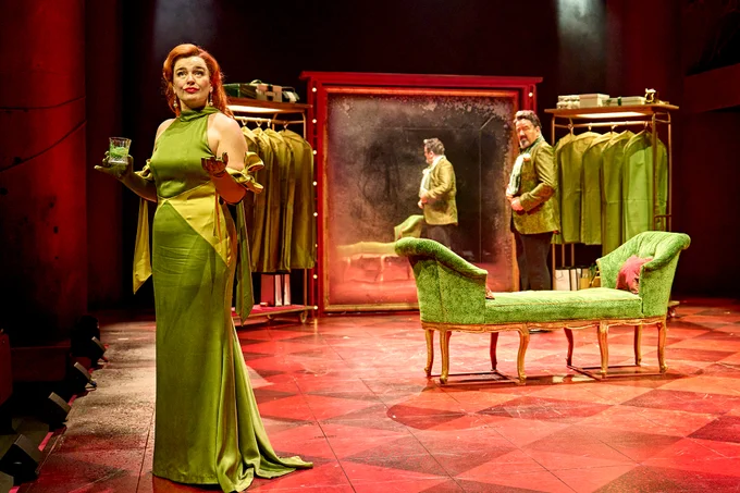 Dianne Pilkington as Mariella stands in a light green silk dress, holding a cocktail. Behind, James Doherty as Bob gets ready in a large mirror. Either side, clothes hang in clothes bags, and in the centre is a large green velvet chaise longue. 