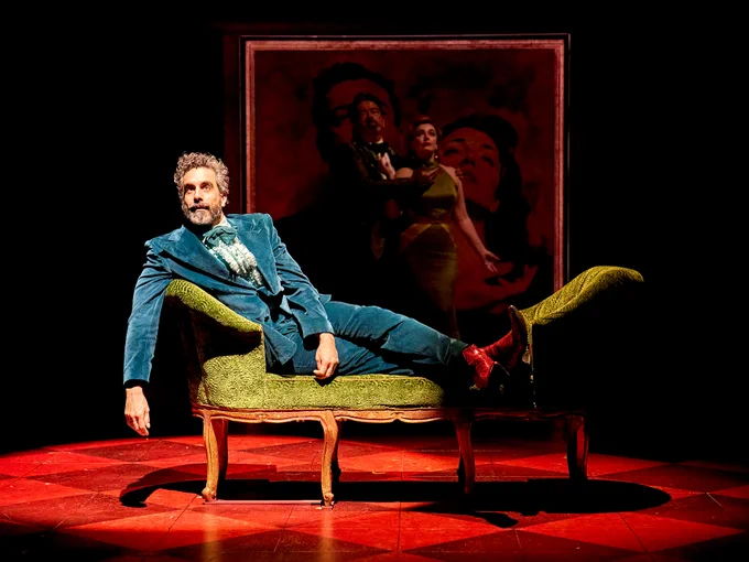 Sebastien Torkia as Sylvestor lies on a chaise longue wearing a blue-grey suit and bow tie. Behind, partially obscured in the dark, are James Doherty as Bob and Dianne Pilkington as Mariella. In the very background is a large red painting of Bob and Mariella