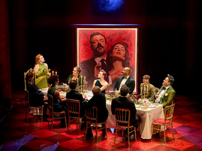The Red Shoes company sit around a large rectangular dining table, eating a meal. Behind there is a large portrait of two people. The floor is checked red.