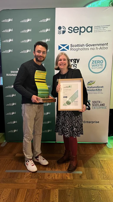 Two people - Elaine Brown -CEO of Edinburgh Remakery and Asrar Qazi, Project & Marketing Executive from Edinburgh Remakery are standing in front of a sponsorship board holding the VIBES award and framed certificate
