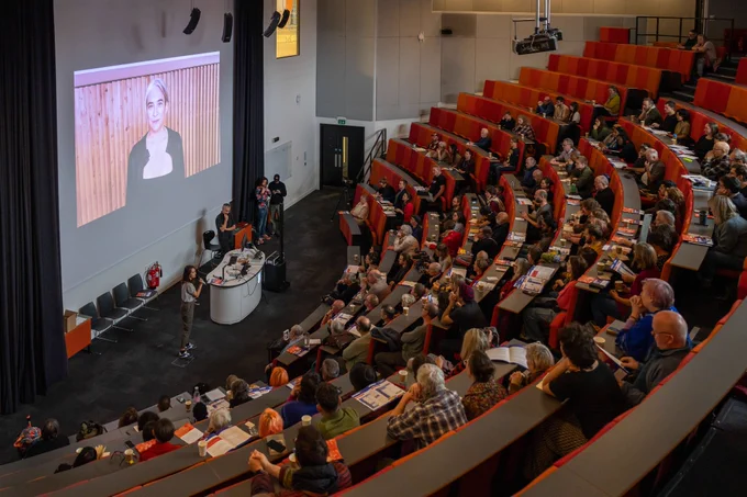 A lecture theatre with bright orange seats, full of people. Standing at the front of the room are a number of people, one of whom is addressing the audience through a microphone. 