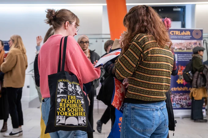 Two people looking at a brochure for the Fearless Cities South Yorkshire summit together. They appear to be discussing the brochure. The person on the left is carrying a black tote bag over their shoulder that reads 'The People's Republic of South Yorkshire'.