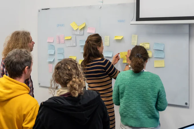 Five people stand in front of a whiteboard covered in pastel-coloured sticky notes. One of the people is placing a sticky note on the whiteboard.