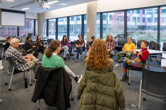 A group of twelve people sit on chairs in a circle, in the middle of a discussion. In the background are a number of large windows, through which buildings outside are visible.