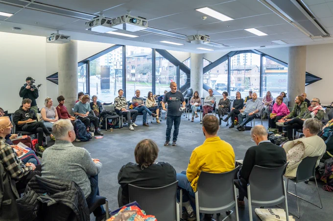 Many people are seated on chairs that have been arranged into a circle. At the centre of the circle, the speaker (Dr Simon Duffy) is talking to the group.