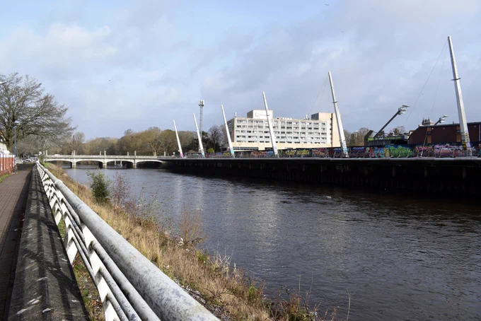 The River Taff from further down next to the Millenium Stadium's walkway, with graffitied walls alongside it and scruffy banks of vegetation on the edges. 