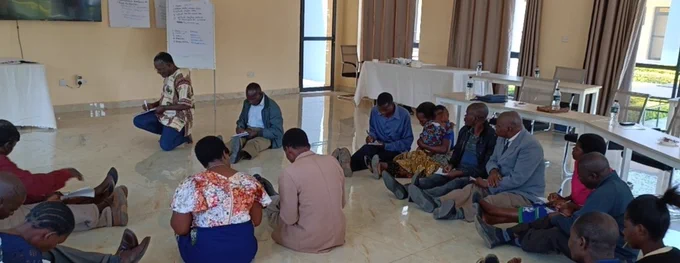 Men and women in a classroom. They are sitting on the floor and they are writing in their notebooks. Behind them are their desks, which are placed in a semi-circle. In the front of the classroom on the wall is a television screen and two pieces of paper taped to the wall. There's an easel with writing on paper and a table against the wall.