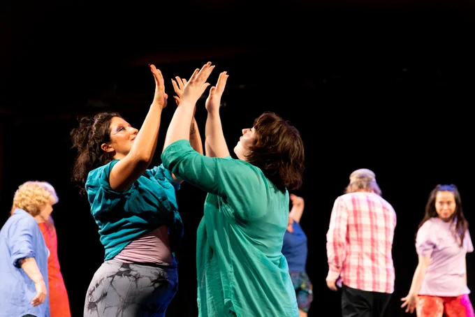 Two dancers dressed in green stand with their forearms raised, almost touching.
