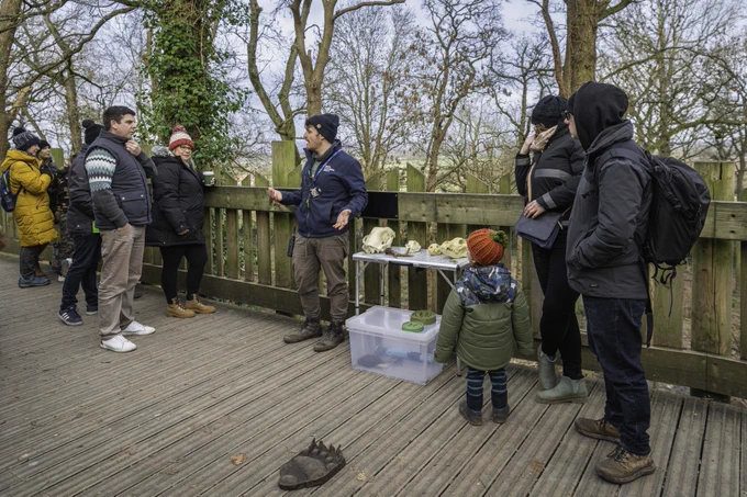 A group of people gather around an educator at an outdoor nature class, discussing animal skulls displayed on a table. Wintertime attire is visible, and the setting includes leafless trees and a wooden deck.