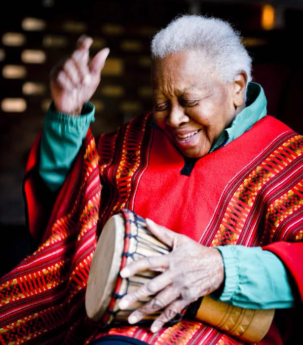 photo of Ella Jenkins, an older Black woman with short gray hair. She wears a red poncho and is playing a djembe drum
