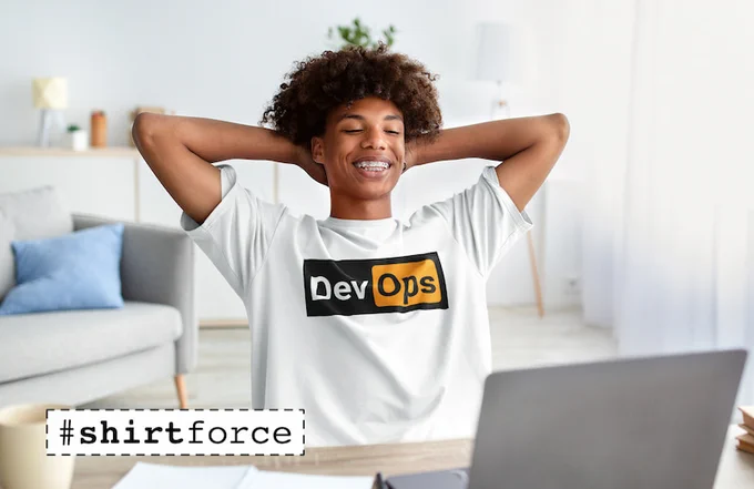 Young man sitting on a chair on front of a laptop. He's smilng and wearing a white T-Shirt with the words DevOps on it. There is a black background beind the whole word, and an orange background behind the "Ops" part.