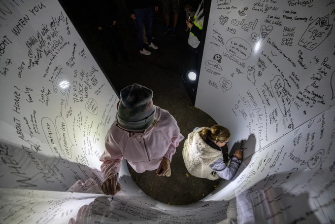 Photograph from above of two people writing on a large curved white wall covered in writing.