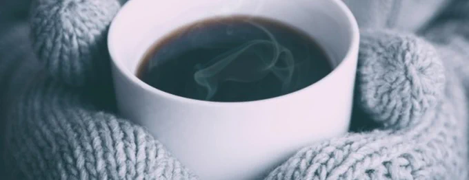 Blue grey mittens hold a steaming white cup of coffee

