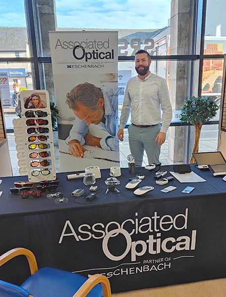 Image is John from Associated Optical standing behind a desk with lots of products and promotional information