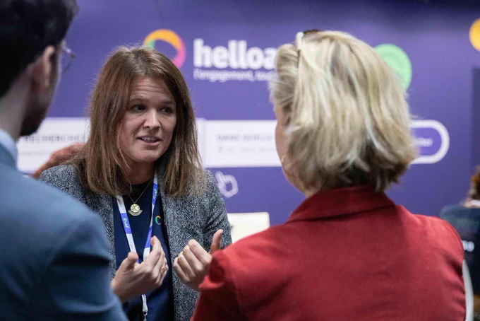 La ministre, de dos, échange avec un membre de l’association « HelloAsso » une entreprise solidaire d’utilité sociale qui propose des outils en ligne pour faciliter la gestion et le développement des associations en France. En arrière plan, un fond violet et le logo de l’association.