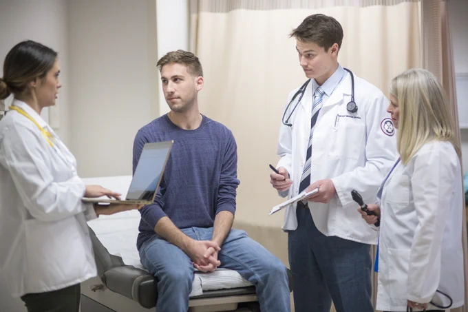 A patient speaks is seated at a hospital bed and speaks to three RFU members in white coats. One of them is holding a laptop.