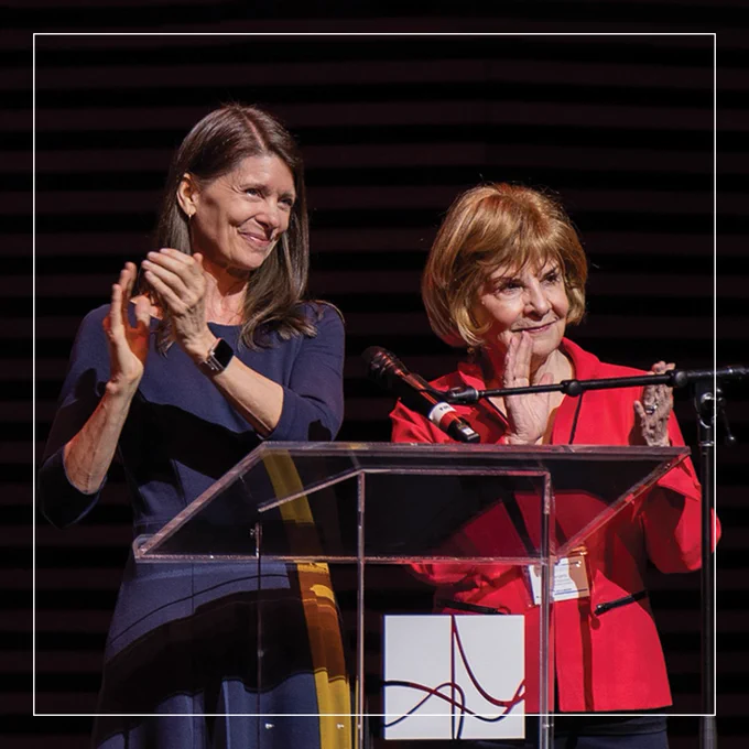 A photograph of two women standing side by side at a podium. They looking out into an audience and smiling as they a round of applause. The women pictured are UF Center for Arts in Medicine's director of research initiatives, Jill Sonke, and Central Florida philanthropist, Margery Pabst Steinmetz, at the Creating Healthy Communities convening.