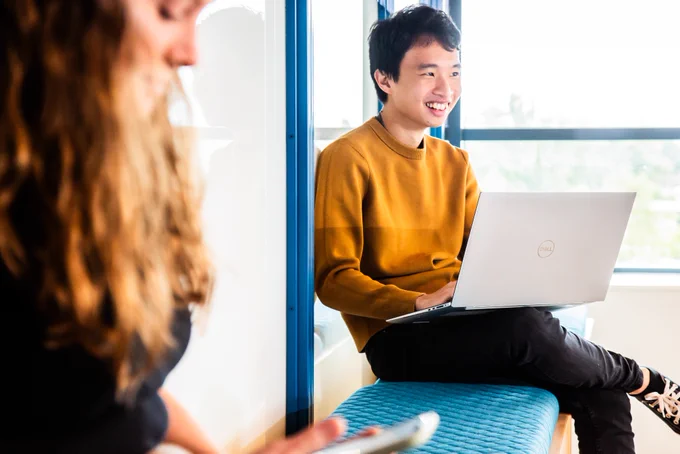 Young engineer sits amongst other people with their laptop. They are laughing at something off screen.