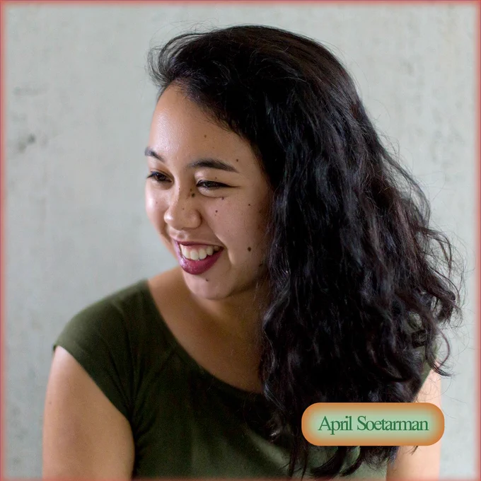 April, an Indonesian-American woman with long dark curly hair, is smiling while looking to the side off-camera. She is wearing a green top in front of a grey cement background.