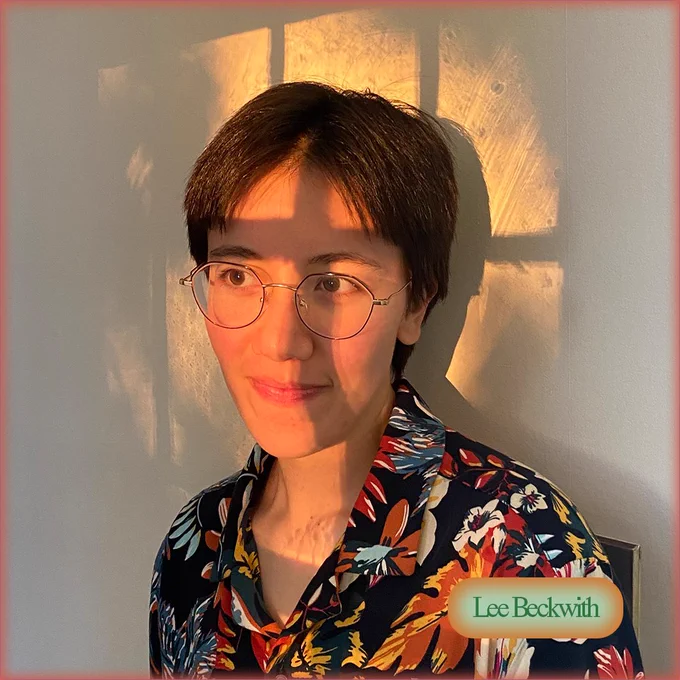 Lee, a white-passing person with short brown hair and wire glasses, stands in a swath of yellow-orange light that has filtered through a window. They wear a navy collared shirt with colorful leaves and flowers on it.